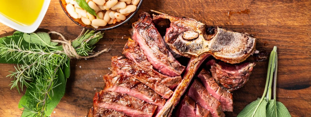 auf einem Holzbrett liegt ein bereits gegrilltes und geschnittenes Bistecca alla Fiorentina, daneben liegen ein Bündel mit frischen Kräutern, eine Schale mit gerösteten Pinienkernen und ein Schälchen mit Olivenöl