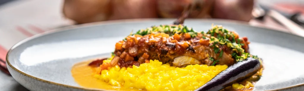 close up von Ossobuco alla Milanese mit Safranrisotto und glasierten Möhren serviert auf einem grauen Steinzeugteller