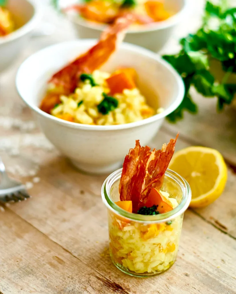 auf einem rustikalen Tisch steht ein kleines Einweckglas, in dem sich Kürbisrisotto befindet. Dieses wurde mit zwei Würfeln vom Kürbis und einem Streifen gebratenen Schinken garniert. Im Hintergrund erkennt man eine Zitrone und Petersilienblätter.