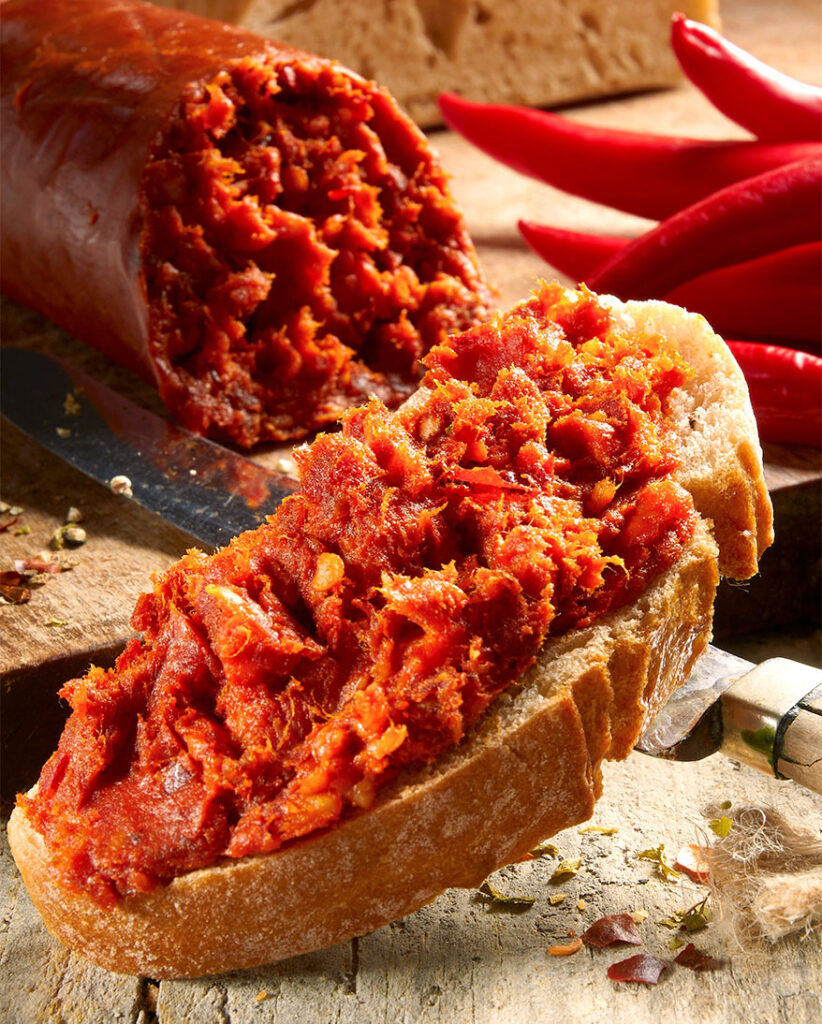zu sehen ist eine frisch aufgeschnittene Nduja am Stück, auf einer Scheibe Ciabatta im Vordergrund ist die Streichsalami bereits verteilt.