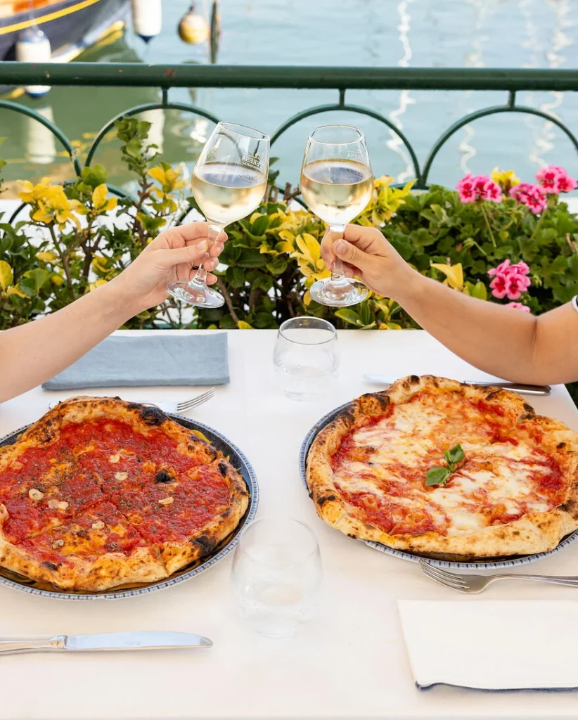 auf einem Tisch eines italienischen Restaurants stehen zwei Teller mit verschiedenen Pizzen: links eine Pizza Marinara und rechts eine pizza Magherita. Es sind weiterhin zwei Arme an den gegenüberliegenden Seiten des Tisches zu sehen, die jeweils ein Glas mit Weißwein in der Hand halten und sich zuprosten.