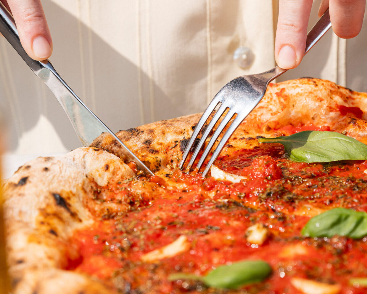 Nahaufnahme: in der Mittagssonne Neapels wird eine frisch gebackene Pizza Marinara mit Messer und Gabel geschnitten