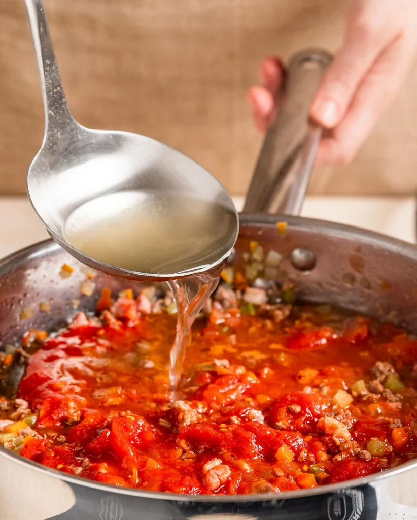 Nahaufnahme eines Kochtopfs in dem ein Ragu gekocht wird, zu sehen ist eine kelle, die Brühe hineingibt, Soffritto, Tomaten und Fleisch sind schon im Topf