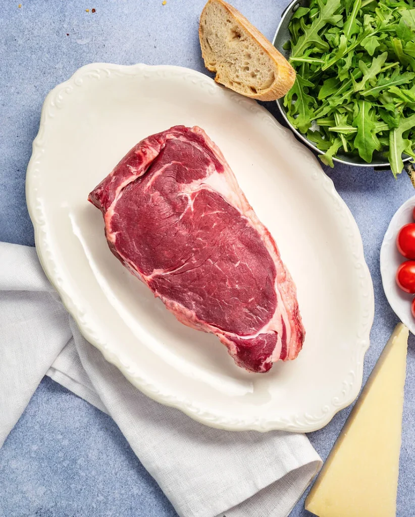 ein rohes Entrecote Steak liegt auf einem weißen Teller, daneben befinden sich Schalen mit Rucola, Kirschtomaten, eine Scheibe Ciabatta und ein Stück Parmiggiano