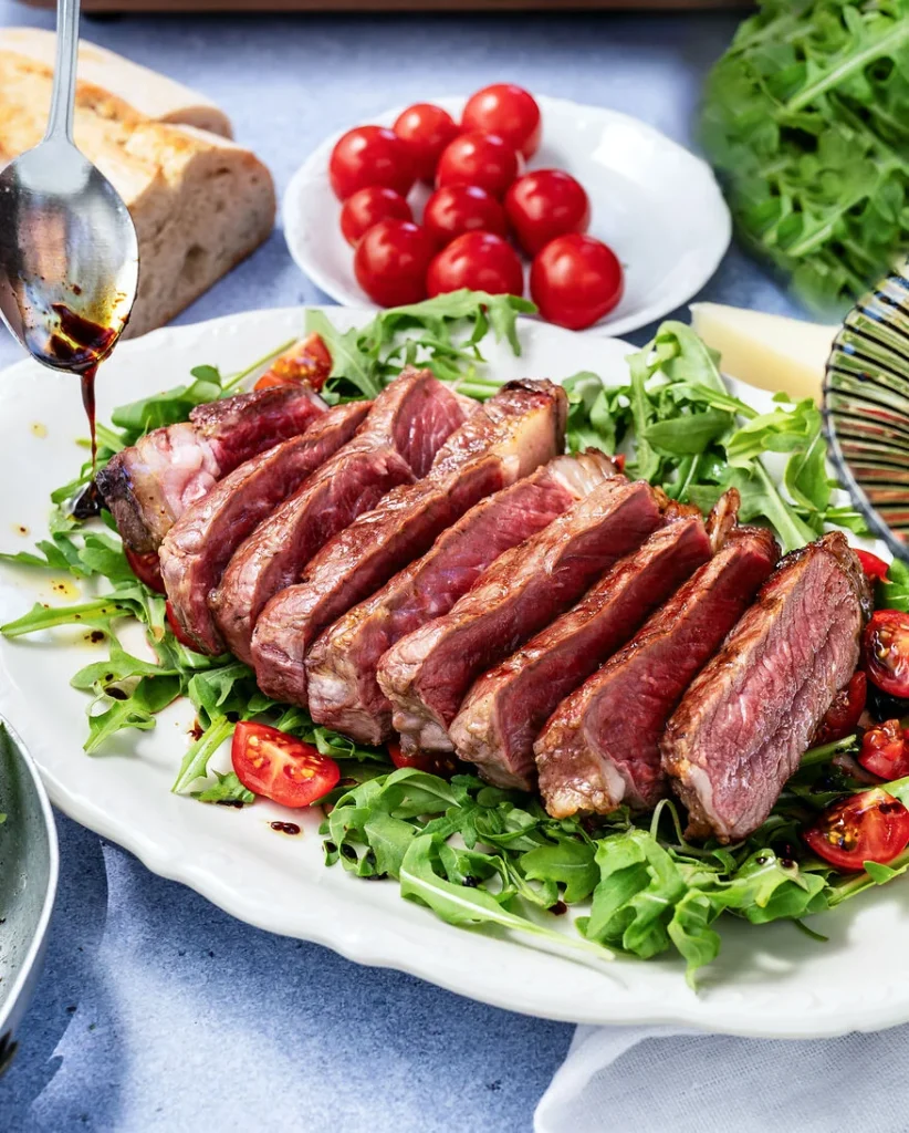 ein fertig gebratenes und bereits geschnittenes Tagliata liegt auf einem Bet von Rucolua und Kirschtomaten, im Hintergrund sind ein Teller mit weiteren Kirschtomaten und ein Stück Ciabatta zu erkennen, ein Löffel träufelt etwas Öl und Balsamico über das Gericht