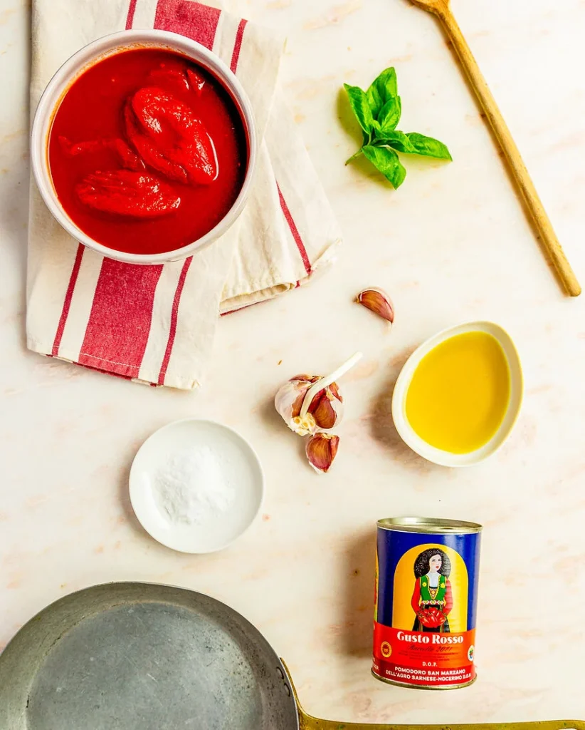 auf einer weißen Marmorarbeitsfläche liegen die Grundzutaten für ein klassisches Sugo: Dosentomaten, Olivenöl, Knoblauch, Salz und Basilikum. Zusätzlich noch die benötigten Kochutensilien: eine Pfanne und ein Kochlöffel