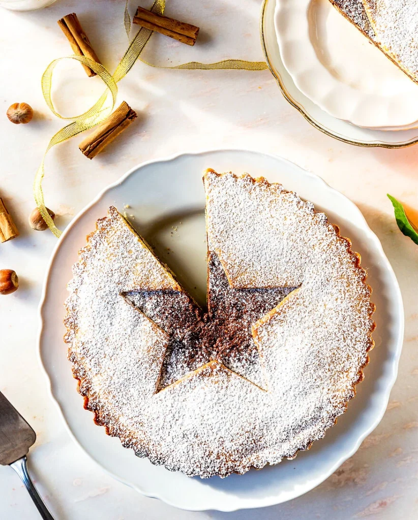 eine Nuss-Tarte mit Stern in der Mitte, ist mit Puderzucker bestreut und liegt in der Mitte eines Tellers, ringsum liegen Zimtstangen und Haselnüsse sowie ein goldenes Geschenkband.