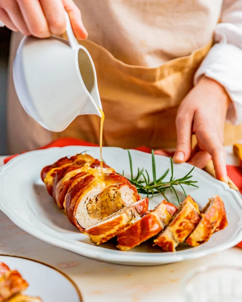 Tacchino im Schinkenmantel: ein moderner Hauptgang bestehend aus gerollter Putenbrust im Schinkenmantel wird auf einem weißen Teller mit Soße übergossen, daneben liegt ein Zweig Rosmarin