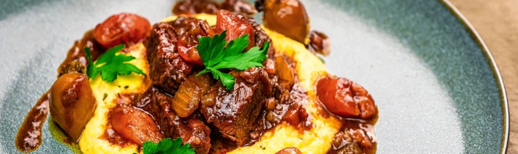 Close up: Spezzatino di Manzo – italienisches Gulasch auf einem blauen Steinzeug Teller angerichtet mit Polenta. Im Hintergrund steht ein Rotweinglas und eine Pfeffermühle