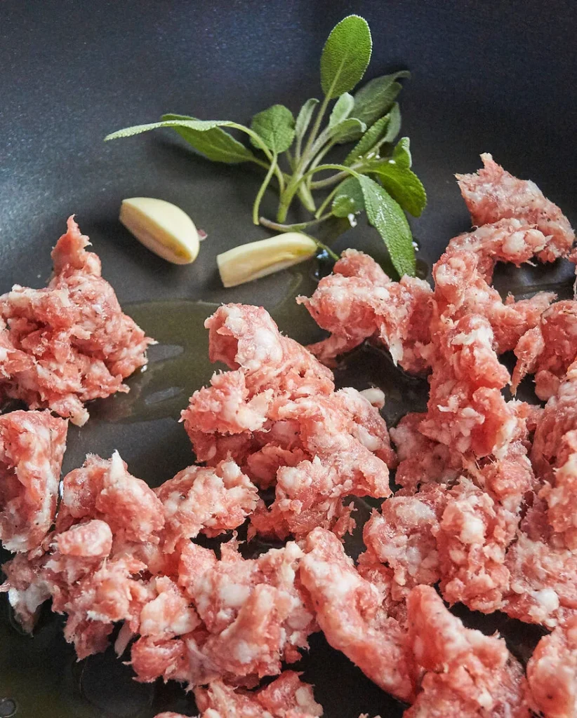 Salsiccia wird in einer Pfanne mit Öl, Knoblauch und Salbei angebraten
