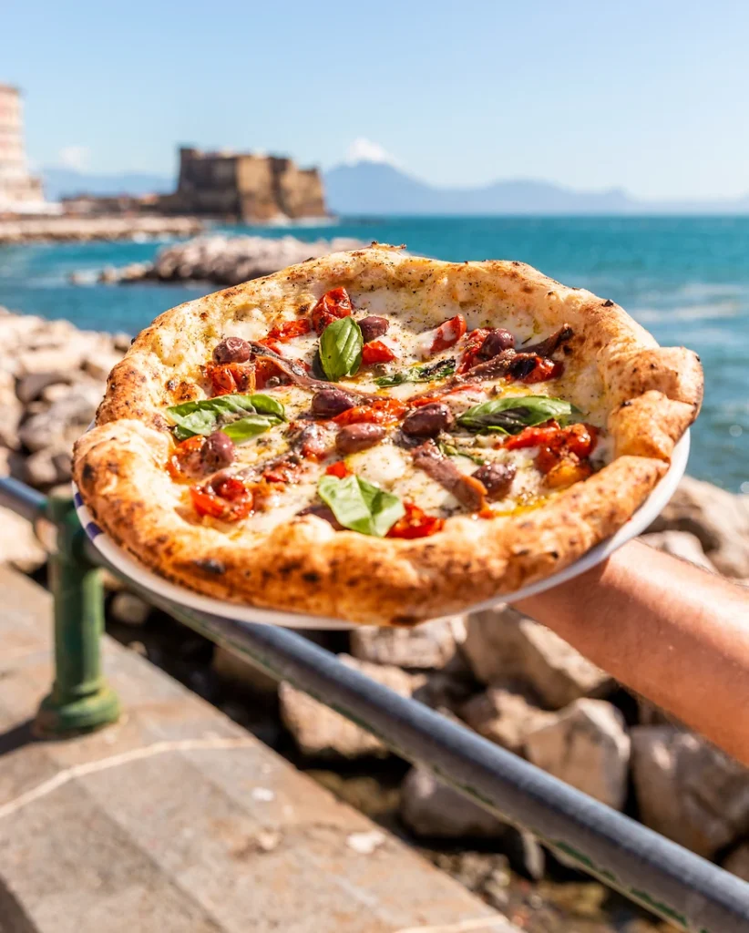 eine Hand hält eine frische Pizza Puttanesca in den Händen vor dem Hafen von Neapel