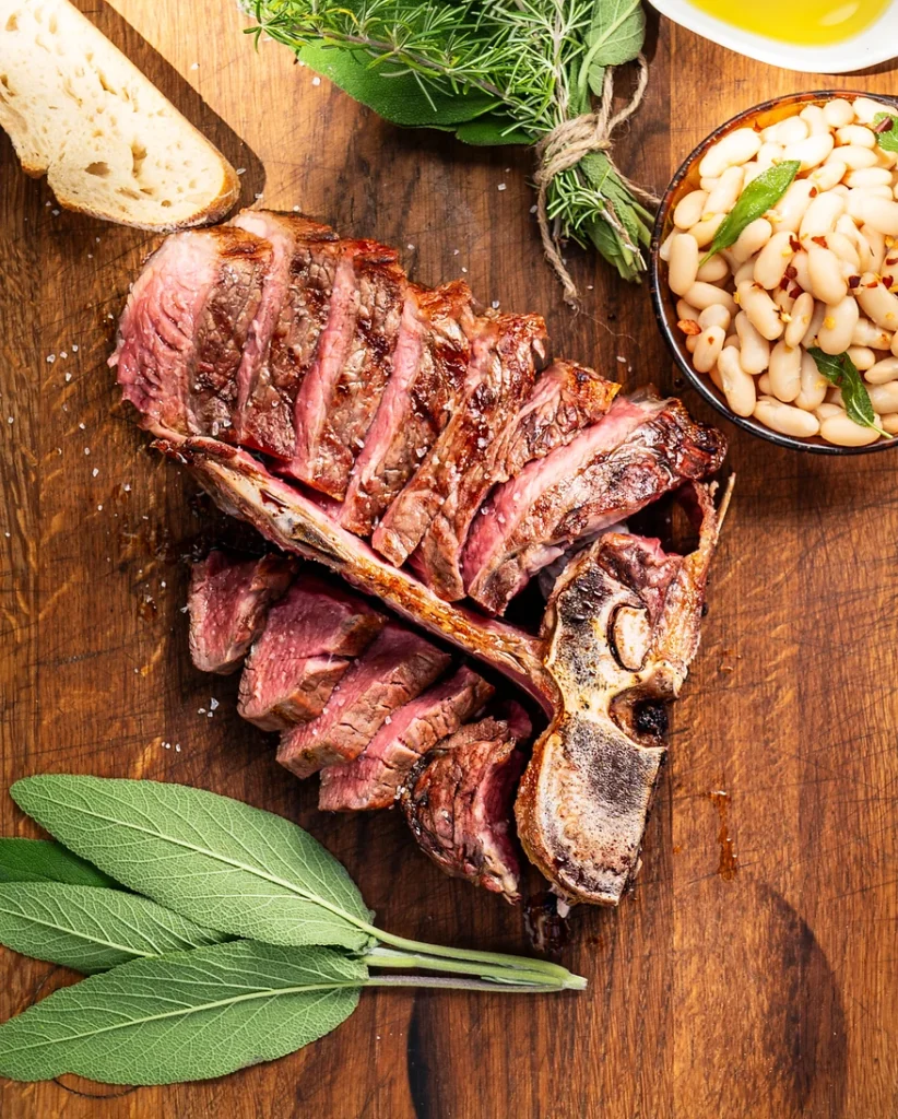Ein fertig gegrilltes Bistecca Chianina liegt auf einem Holzbrettchen, bereits in Scheiben geschnitten, umringt von einem Bündel Salbei, Pinienkernen und Olivenöl