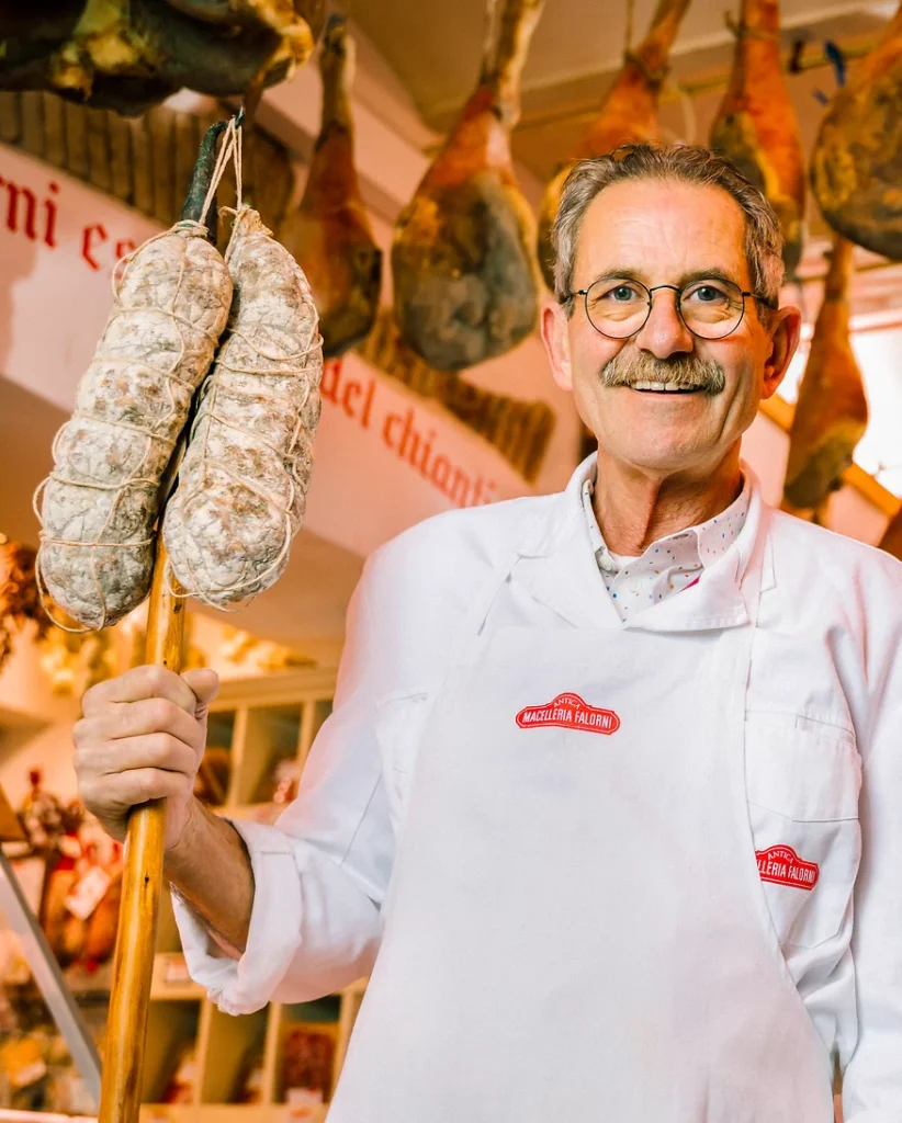Stefano Falorni steht in seiner Macelleria mit zwei großen reifen Salamis und lächelt in die Kamera