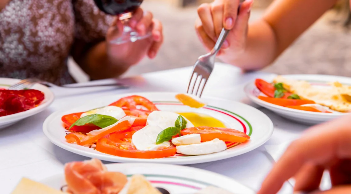 In einer Gasse Italiens steht ein Tisch gedeckt mit verschiedenen Antipastiplatten, im Fokus ist ein Teller mit Caprese zu sehen, eine Frau ist gerade dabei mit einer Gabel eine Scheibe Tomate zu nehmen
