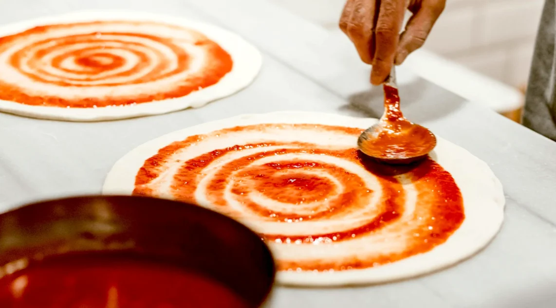 auf einer weißen Arbeitsfläche werden zwei Pizzateige in kreisförmigen Bewegungen mit Soße bestrichen