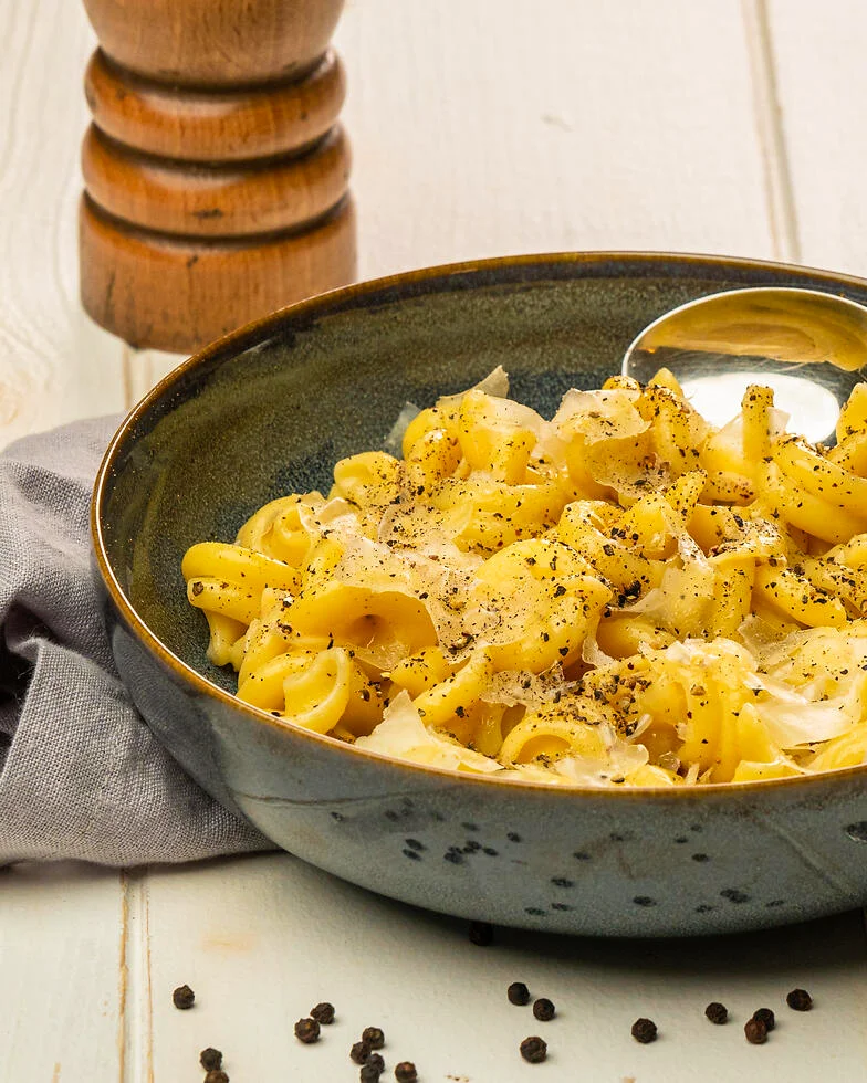 auf einem weißen Tisch steht ein blauer Steinzeug-Teller mit frisch angerichteten Pasta Vesuvio Cacio e Pepe. Im Vordergrund liegen einige Pfefferkörner vor dem Teller, dahinter steht die Pfeffermühle