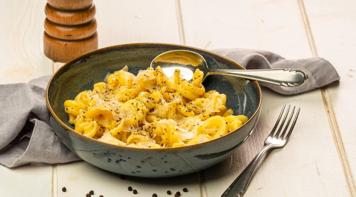 pasta cacio e pepe serviert auf einem blauen Steinzeug-Teller, dieser steht auf einem weißen Tisch eine Pfeffermühle steht im Hintergrund
