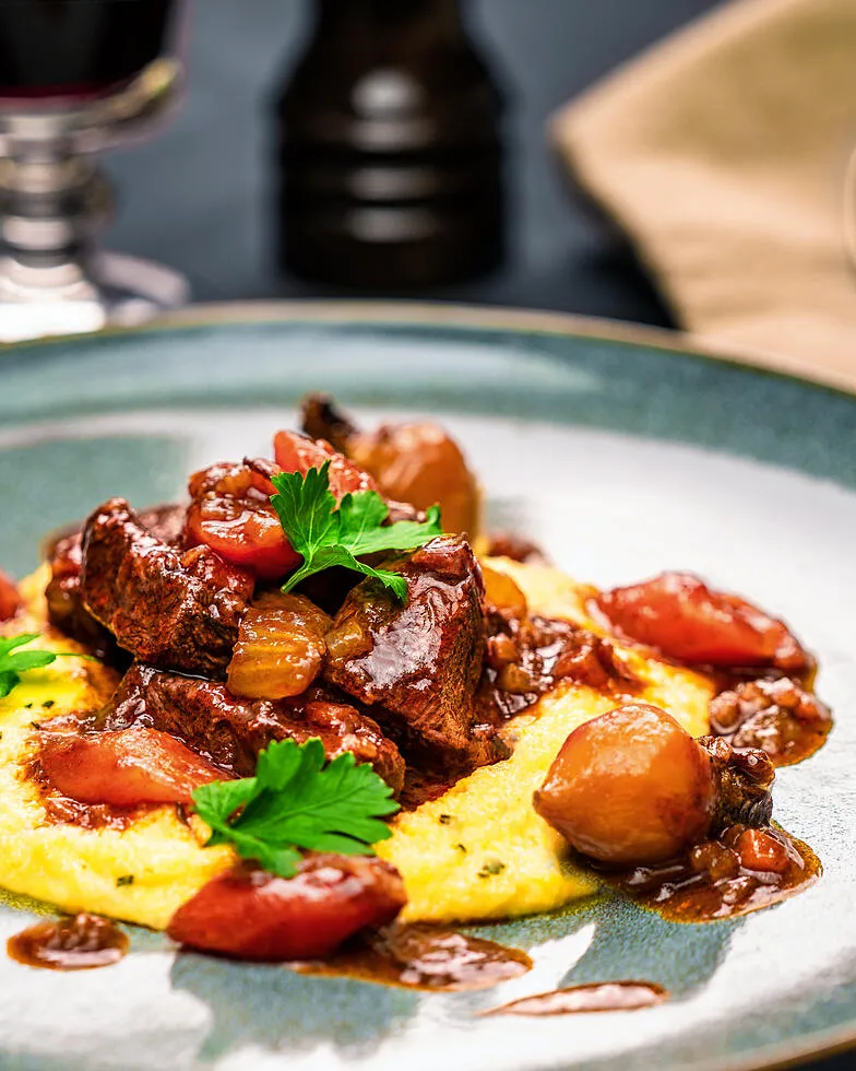 Spezzatino di Manzo – italienisches Gulasch auf einem blauen Steinzeug Teller angerichtet mit Polenta. Im Hintergrund steht ein Rotweinglas und eine Pfeffermühle