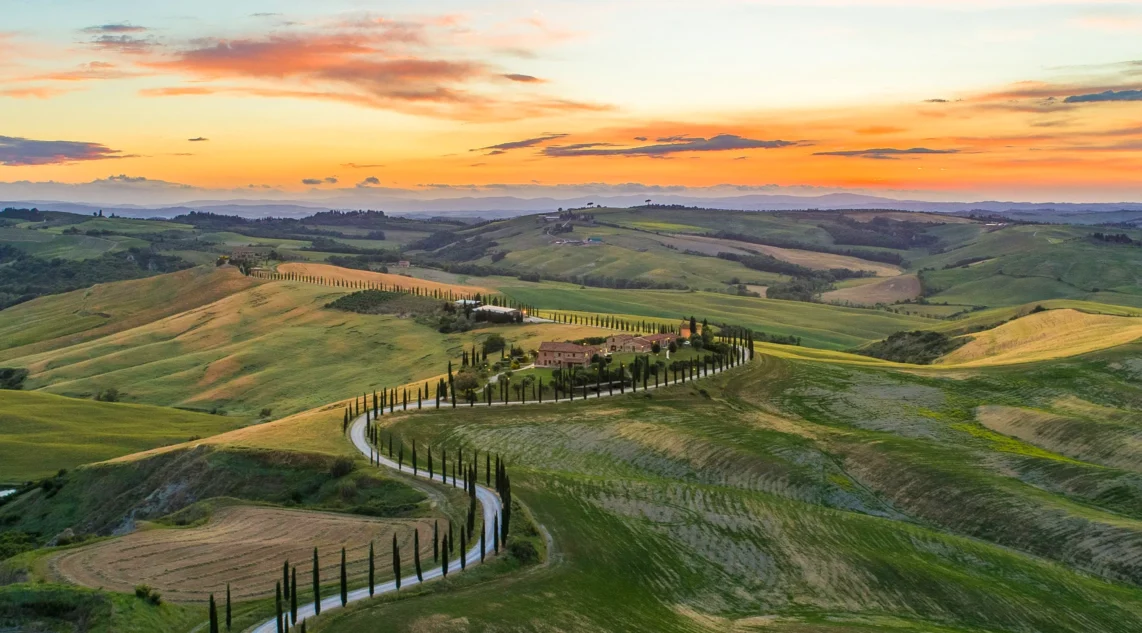Sonnenuntergang im Panoramaformat: Das Valle D'Orcia mit grünen Hügeln, Zypressenbäumen und mediterranen Häusern liegt friedlich unter einem dramatischen Himmel in Orange-Blau-Violett