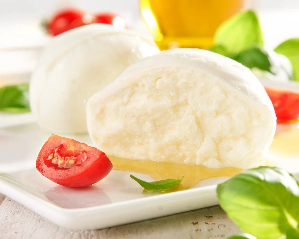 eine angeschnittene Kugel Mozzarella liegt auf einem weißen Teller, im Vordergrund liegt ein Blatt Basilikum und daneben eine Tomate
