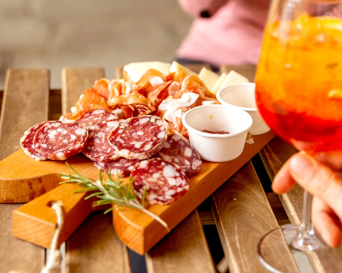 auf einem Tisch liegt eine Antipastiplatte mit Salamischeiben, eine Hand hält einen Aperol Spritz
